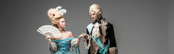 панорамный снимок красивой женщины с веером, смотрящей на джентльмена в парике на сером
