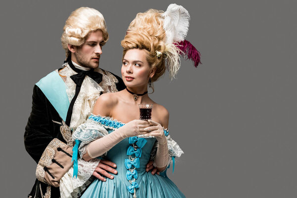 victorian gentleman in wig looking at beautiful woman with wine glass isolated on grey 