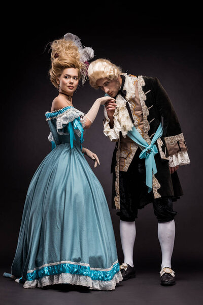 Красивый викторианский мужчина целует руку женщины в парике, стоя на черном
 