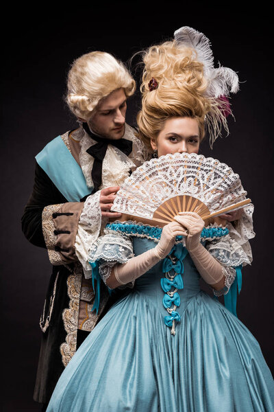 handsome man looking at victorian woman in wig covering face with fan on black 