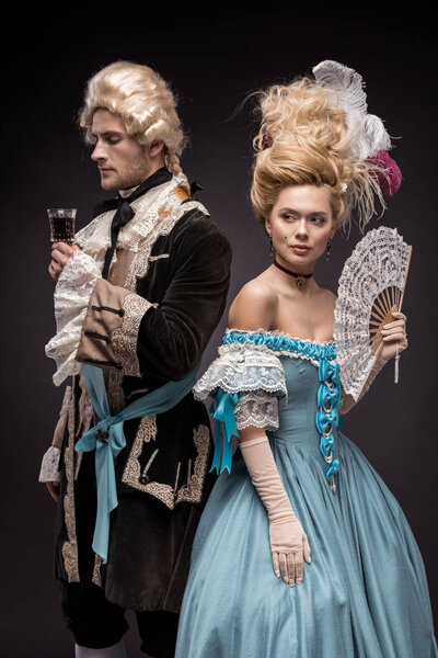 handsome man with wine glass near victorian woman in wig holding fan on black 