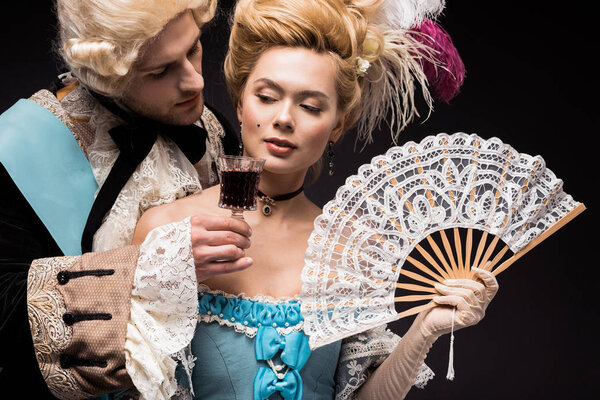 handsome man looking at young victorian woman in wig holding fan  and wine glass isolated on black 