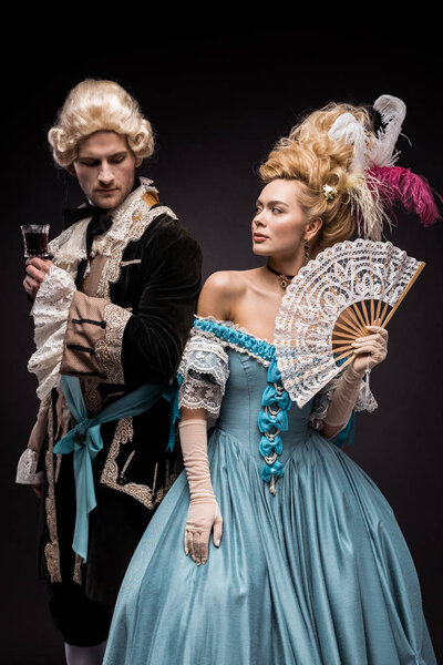 handsome man with wine glass near young victorian woman in wig holding fan on black 