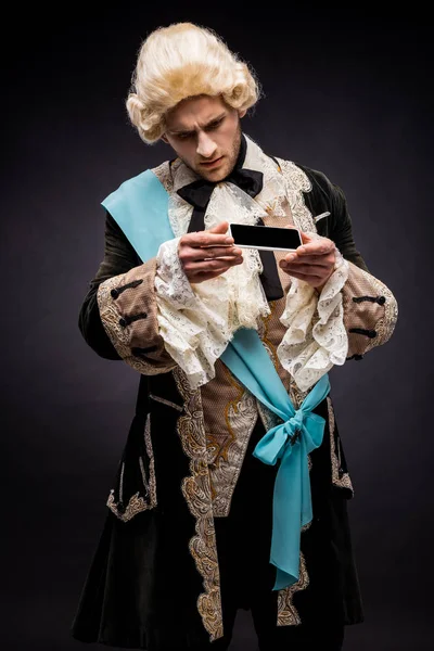 handsome victorian man in wig looking at smartphone with blank screen on black