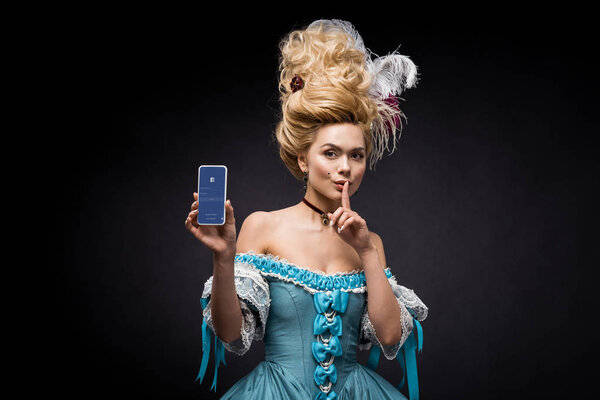 KYIV, UKRAINE - JUNE 5, 2019: attractive victorian woman holding smartphone with facebook app on screen and showing hush sign on black 