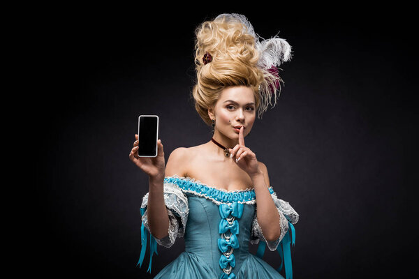 young victorian woman in blue dress holding smartphone with blank screen and showing hush sign on black 