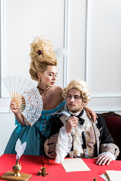 beautiful victorian woman holding fan and standing near pompous man holding feather near papers 