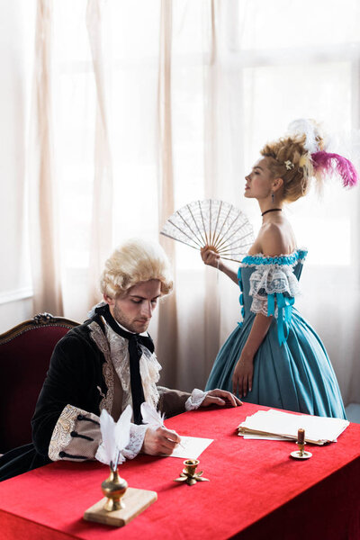 victorian woman in wig standing and holding fan near handsome man writing letter 