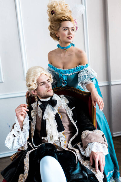 Викторианка, стоящая рядом с мужчиной в парике, сидящая на антикварном стуле
 