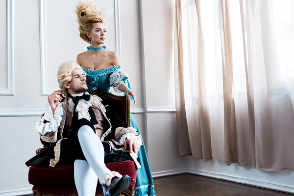 Викторианка, стоящая рядом с красивым мужчиной в парике, сидит на антикварном стуле
 