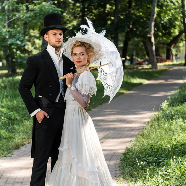 handsome victorian man standing with hand in pocket near woman holding umbrella