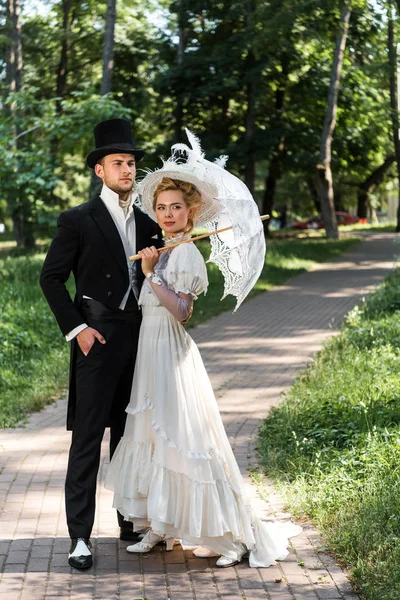 handsome victorian man standing with hand in pocket near woman holding umbrella