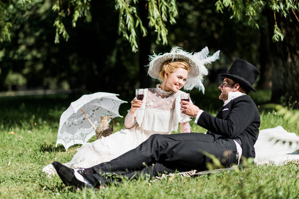 счастливые мужчина и женщина, сидящие на одеяле и держащие в руках бокалы вина
 