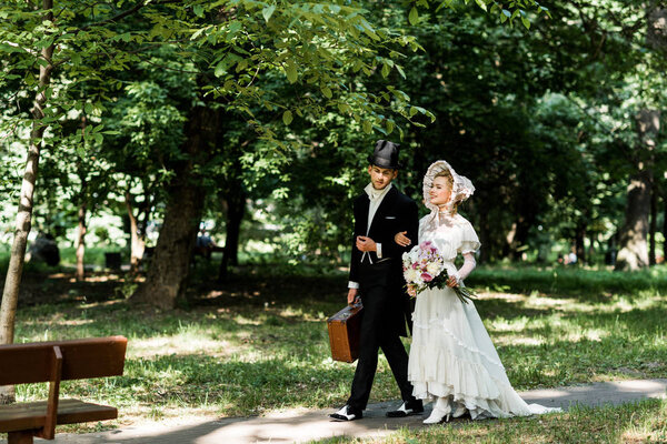 cheerful victorian woman with flowers walking near man with baggage 