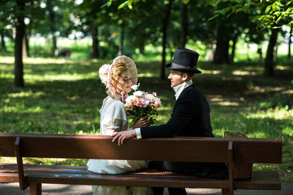 handsome aristocratic man sitting with cheerful victorian woman in hat on bench 