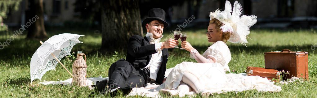 panoramic shot of happy man and woman siting on blanket and clinking wine glasses 