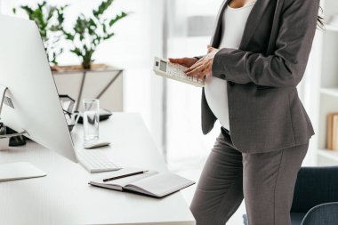 cropped view of pregnant woman calculating while standing near table with computer and notebook in office clipart