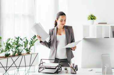  bilgisayar, belge tepsisi ve kalem kutusu ile saksı ve masa yakınında ofiste dururken kağıtları okuma hamile kadın