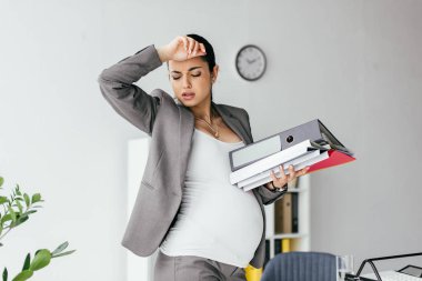 exhausted pregnant woman enduring pain and holding folders  clipart