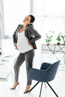 full length view of pregnant woman standing near office chair in office and enduring pain clipart