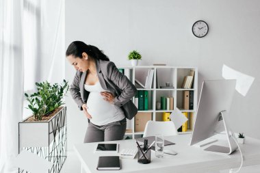 exhausted pregnant woman standing in office and enduring pain clipart