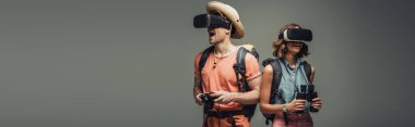 panoramic shot of two young tourists using virtual reality headsets on grey background clipart