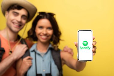 Kiev, Ukrayna - 3 Haziran 2019: Neşeli genç kadının erkek arkadaşının yanında durup sarı arka planda Spotify uygulamasıyla akıllı telefonu göstermesinin seçici odağı.