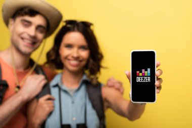 Kiev, Ukrayna - 3 Haziran 2019: Neşeli genç kadının erkek arkadaşının yanında durup sarı arka planda Deezer uygulamasıyla akıllı telefonu göstermesinin seçici odağı.