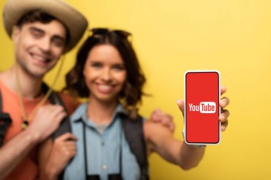 Kiev, Ukrayna - 3 Haziran 2019: Neşeli genç kadının erkek arkadaşının yanında durup youtube uygulamasıyla sarı arka planda akıllı telefonu göstermesinin seçici odağı.