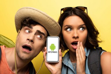 Kiev, Ukrayna - 3 Haziran 2019: Sarı arka plan üzerinde şok adam yanında dururken Android uygulaması ile akıllı telefon gösteren sürpriz genç kadın.