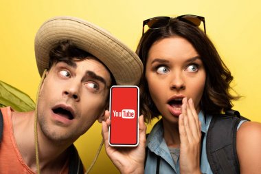 Kiev, Ukrayna - 3 Haziran 2019: Sarı arka planda şok bir adamın yanında dururken Youtube uygulamasıyla akıllı telefon gösteren genç kadını şaşırttı.