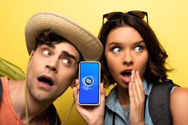 Kiev, Ukrayna - 3 Haziran 2019: Shazam uygulaması ile akıllı telefon gösteren şaşıran genç kadın sarı arka planda şok adam yanında dururken.