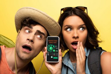 shocked young woman showing smartphone with marketing analysis app while standing near surprised man on yellow background clipart