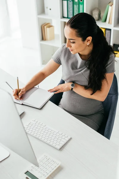 孕妇坐在白色桌子后面 电脑和键盘 在笔记本上做笔记的高角度视图 — 图库照片