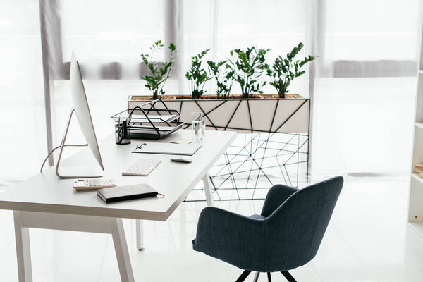 white table with computer, document tray, glass and notebook near office chair and flowerpot with plant