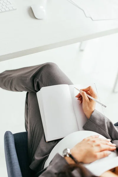 オフィスの椅子に座っている間 ノートにメモを取り 腹を保持している妊娠中の女性をトリミングビュー — ストック写真