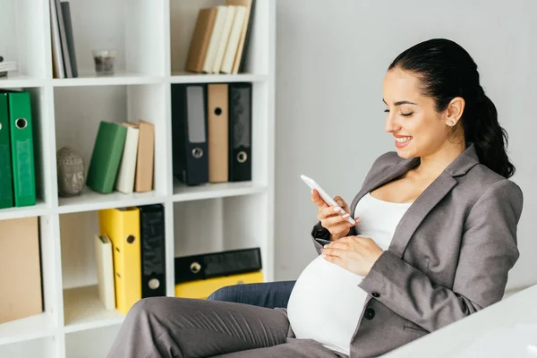 オフィスチェアに座って 笑顔でスマートフォンを見ている妊娠中の女性 — ストック写真