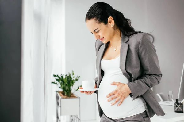 ソファのカップと耐え続けの痛みでオフィスに立っている妊娠中の女性 — ストック写真