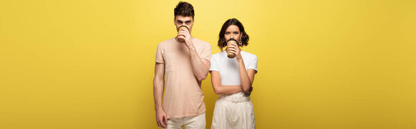 panoramic shot of young man and woman drinking coffee to go while looking at camera on yellow background