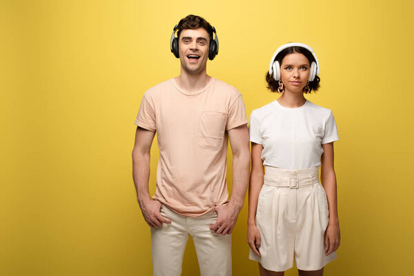 cheerful man and woman posing at camera while listening misic in headphones on yellow background