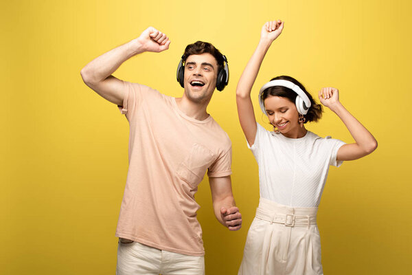 happy young man and woman dancing while listening music with headphones on yellow background