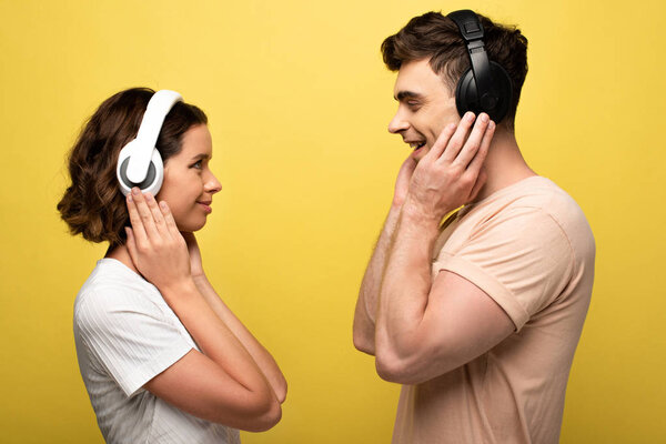 cheerful man and woman listening music in headphones while looking at each other on yellow background