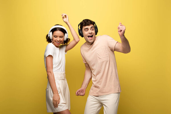 excited man and woman dancing while listening music in headphones on yellow background