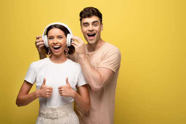 cheerful man putting on headphones on excited girl while looking at camera together on yellow background