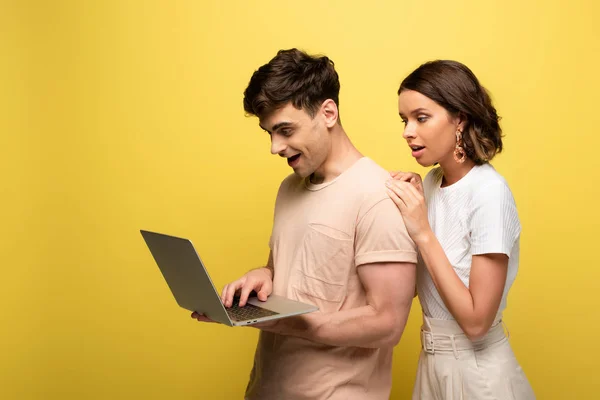 smiling young man using laptop while standing near pretty girl on yellow background