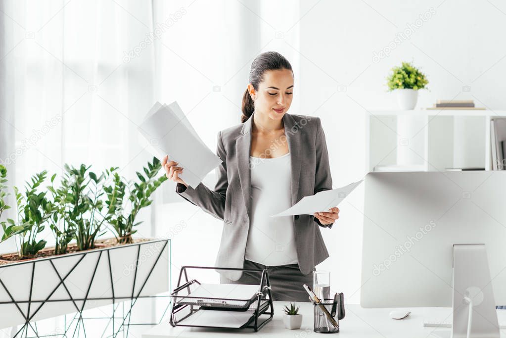  pregnant woman reading papers while standing in office near flowerpot and table with computer, document tray and pencil box