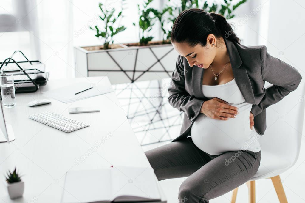 pregnant woman sitting in chair behind table in office, holding belly with hands and enduring pain