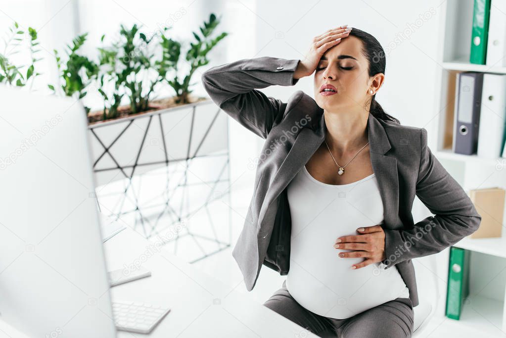 pregnant woman sitting in office behind table and holding head with hand because of pain 