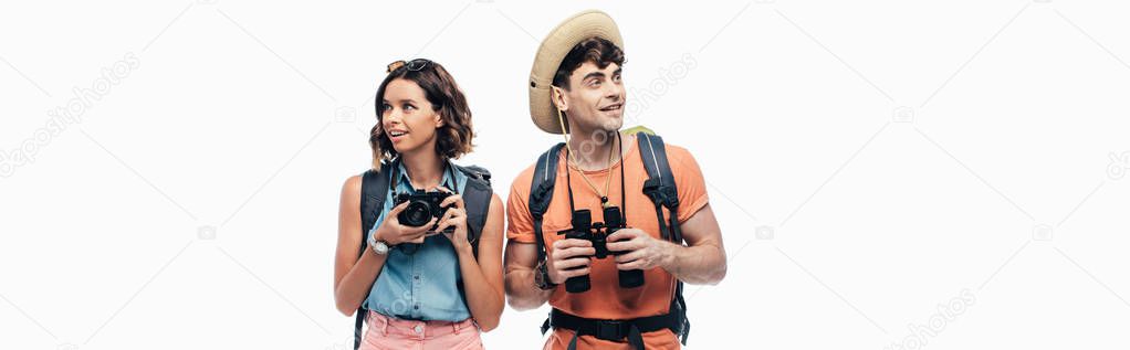 panoramic shot of two smiling tourists with digital camera and binoculars looking away isolated on white