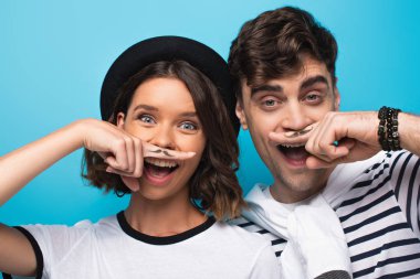 cheerful man and woman holding fingers with drawn mustache near faces on blue background clipart